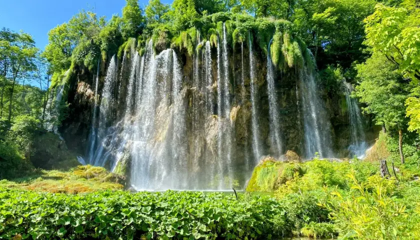 A stunning waterfall cascades down a rocky cliff, surrounded by lush green foliage. Perfect for Croatia hiking trips, this scene captures the natural beauty of Plitvice Lakes National Park.