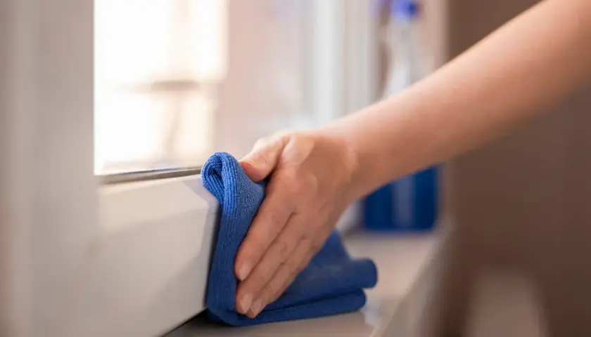 Mano pulisce davanzale con panno blu. Un flacone di detergente è sullo sfondo. Scopri come pulire gli scuri in legno.