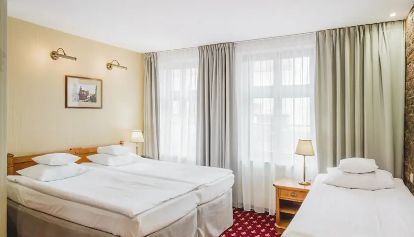 Pokój hotelowy z trzema łóżkami, idealny na wieczór panieński. Jasne ściany, zasłony i czerwony dywan tworzą przytulną atmosferę.