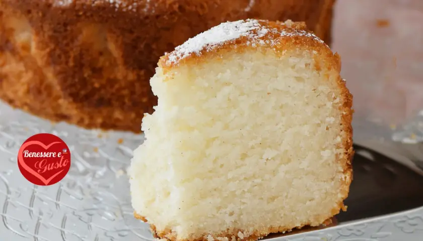 Una fetta di torta senza glutine, soffice e dorata, cosparsa di zucchero a velo, su un piatto trasparente.