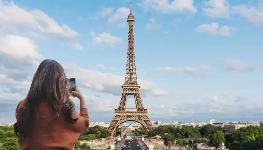 Kobieta robi zdjęcie telefonem ikonicznej wieży Eiffel. Paryski krajobraz z zielenią i budynkami w tle.