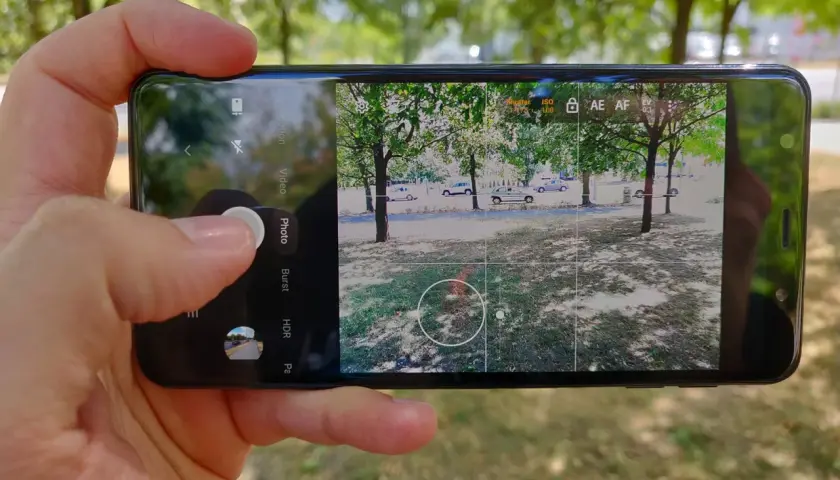 Dłoń trzyma smartfon, na ekranie którego widać interfejs aparatu. Aplikacja aparatu jest gotowa do zrobienia zdjęcia.