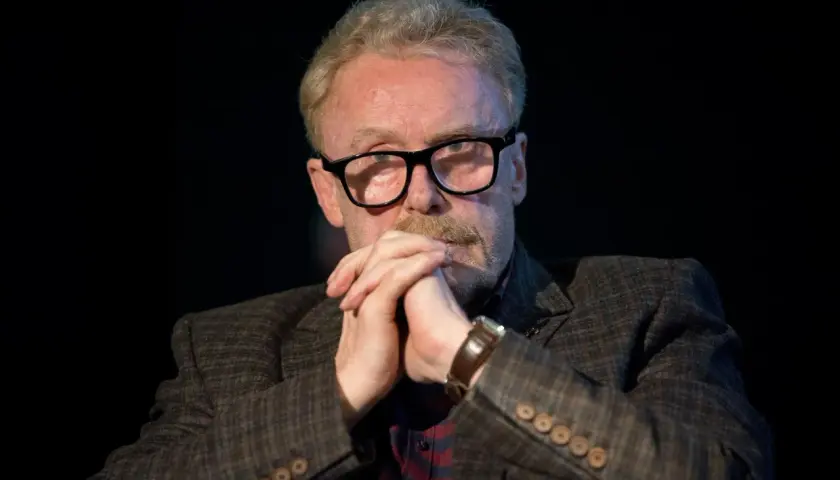 Daniel Olbrychski w okularach, z dłońmi złożonymi na brodzie. Choć nie widać tego na zdjęciu, ile wzrostu ma Daniel Olbrychski, jego postawa sugeruje pewność siebie.