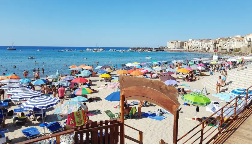 Plage animée à Cefalù en Sicile, avec des parasols colorés, des baigneurs dans la mer bleue et des bâtiments historiques en arrière-plan.