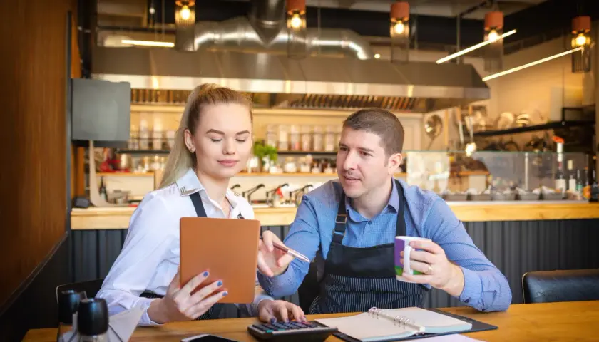 Para omawia koszty uzyskania przychodu w restauracji, analizując dane na tablecie i kalkulatorze.