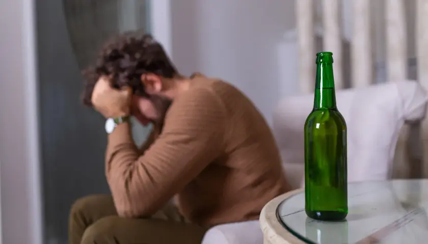 Mężczyzna z głową w dłoniach siedzi obok pustej butelki piwa. Może to symbolizować uczulenie na alkohol lub jego nadużywanie.
