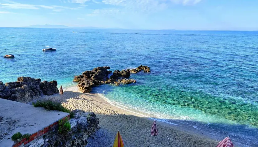 Albania, czerwiec: słoneczna pogoda, turkusowe morze, kamienista plaża z parasolami i łódkami na horyzoncie.