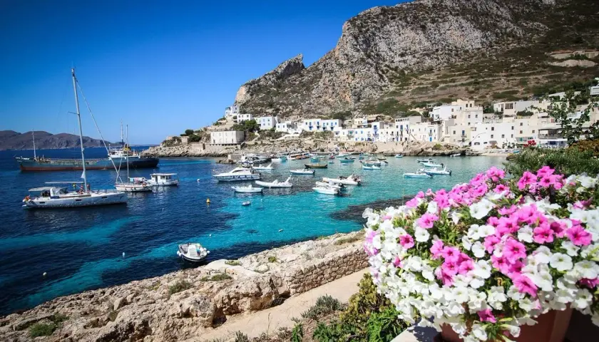 Vue d'un port pittoresque en Italie, avec des bateaux dans des eaux turquoise et des maisons blanches accrochées à flanc de colline. Un bouquet de fleurs roses et blanches encadre la scène, évoquant la beauté de Marsala, Italie.