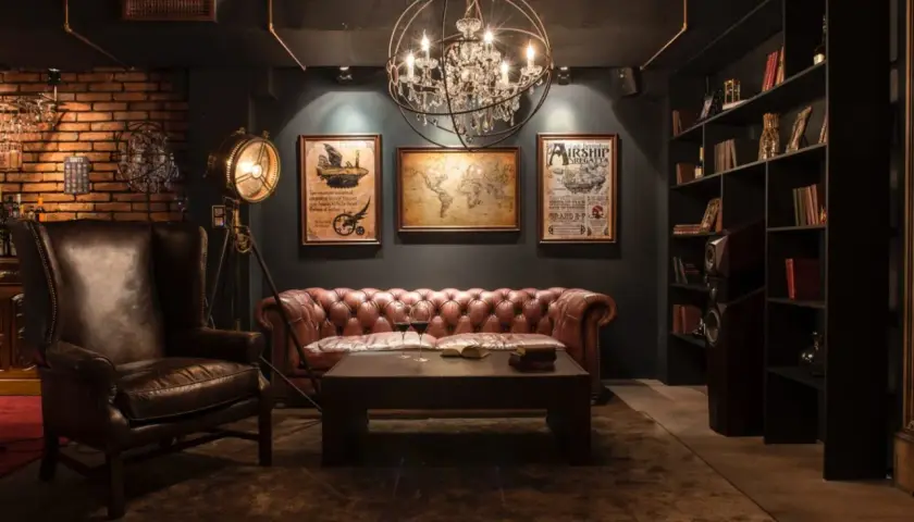 Sofá de cuero, sillón y mapa del mundo en una sala con decoración steampunk. Lámpara de pie industrial y estanterías llenas de libros.