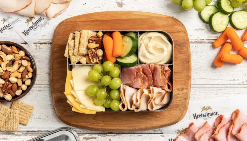 A bento box with cheese, grapes, crackers, nuts, hummus, carrots, and cucumber. Perfect for when are bento boxes eaten cold.
