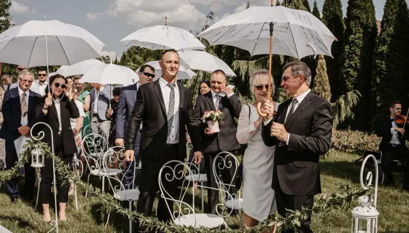 Wesele w kwietniu pod białymi parasolami. Goście w eleganckich strojach czekają na ceremonię na trawie, wśród zieleni i ozdobnych krzeseł.