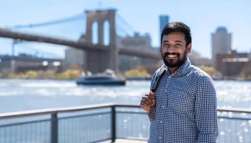 A smiling man with a beard, wearing a blue checkered shirt, stands with the Brooklyn Bridge in the background. He's a student in the Pace University MPA program.