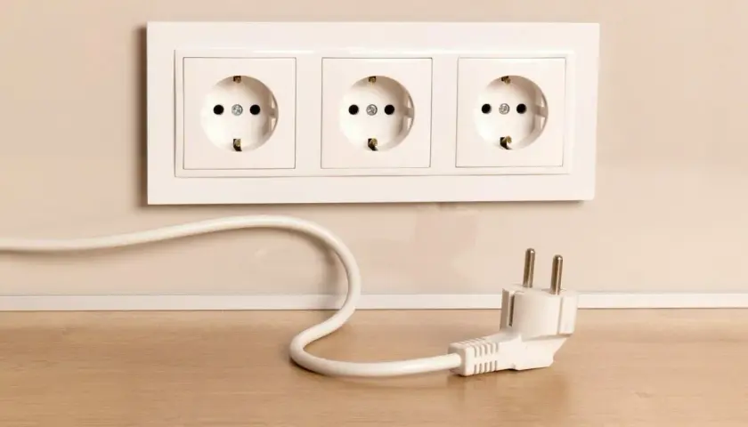 Biały kabel z wtyczką leży na drewnianym blacie, obok gniazdek elektrycznych w ścianie.