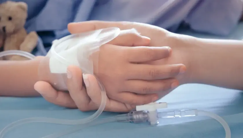 Dłoń dorosłego obejmuje dłoń dziecka z wenflonem. W tle pluszowy miś. Obraz symbolizuje wsparcie i troskę, jakiej wymaga zasilek pielegnacyjny.