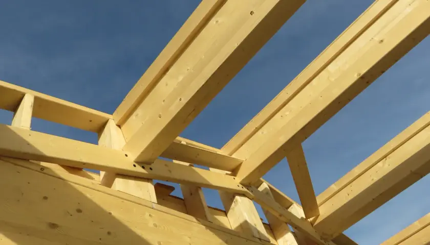 Structure en bois d'une charpente fermette sous un ciel bleu. Les poutres de bois clair forment un motif géométrique complexe.
