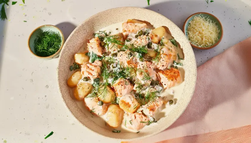 Pyszne gnocchi z łososiem w kremowym sosie, udekorowane koperkiem i kaparami. Obok miseczka z tartym parmezanem.