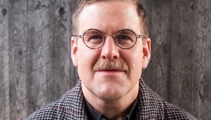Adam Svanell med glasögon och mustasch, klädd i en rutig rock, framför en grå bakgrund.