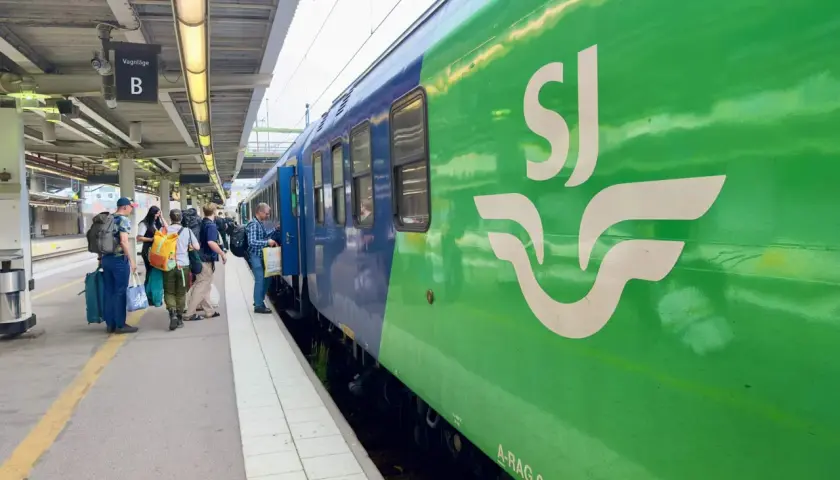 Folk väntar på tåget vid perrong B. En grön SJ-tågvagn syns tydligt. Kanske är det början på en resa till England från Sverige.