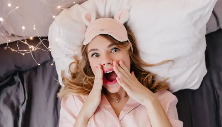 En tjej med sovmask och rosa pyjamas ligger i sängen, redo för att sova med sminket kvar. Hon ser överraskad ut.