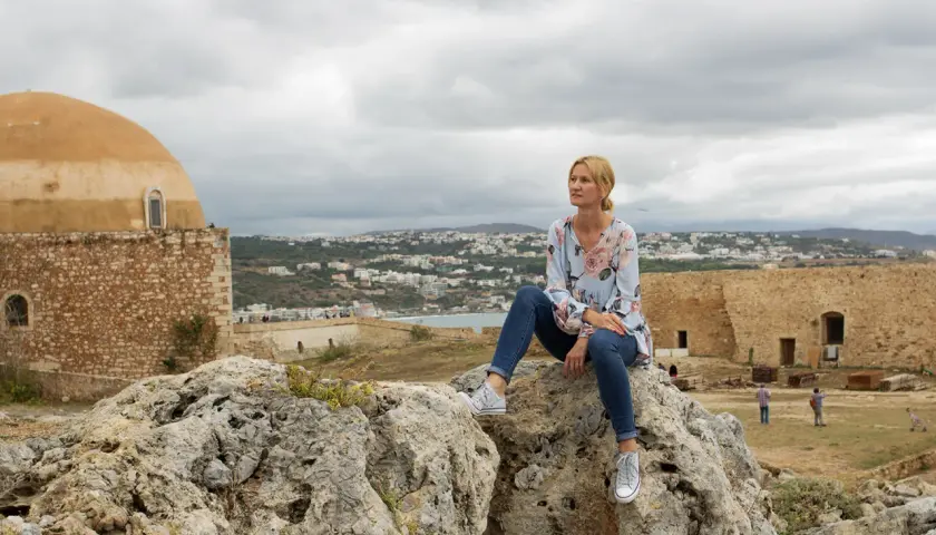 Kobieta siedzi na skale, podziwiając widok miasta. Kreta pogoda październik, chmury na niebie.