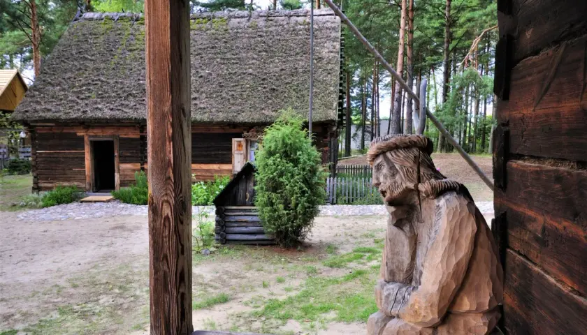 Drewniana rzeźba postaci, tradycyjna chata z mchem na dachu i zielone drzewa tworzą klimatyczne **kadzidło atrakcje** w skansenie.