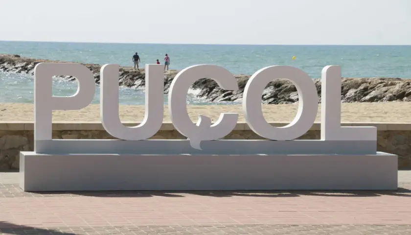 Letras gigantes "PUZOL" en la playa, con el mar azul y personas al fondo.