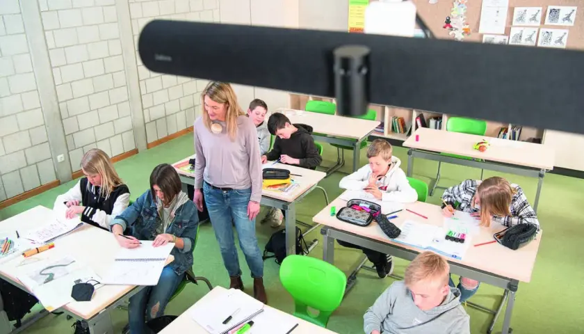 Lehrerin betreut Schüler im Klassenzimmer, die an ihren Tischen arbeiten. Ein modernes Setup für hybriden Unterricht.