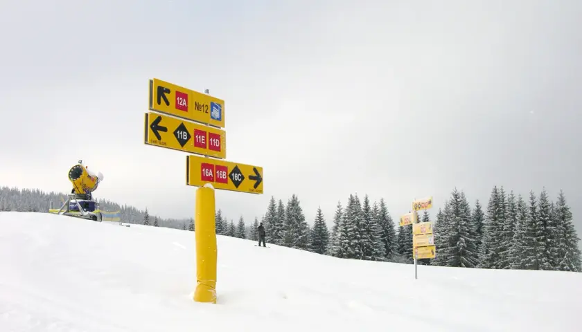 Na stoku narciarskim, wśród zasypanych śniegiem drzew, żółte tabliczki kierują narciarzy na różne trasy.