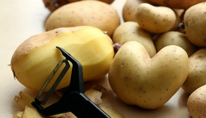 Ziemniaki, bogate w węglowodany, są obierane za pomocą obieraczki. Widać skórki i kilka ziemniaków.