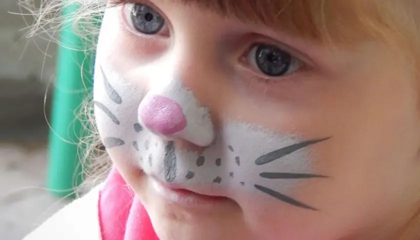 Ein kleines Mädchen mit blauem Auge und Kinderschminken als Tier. Die Nase ist rosa, die Wangen mit Punkten und Schnurrhaaren bemalt.