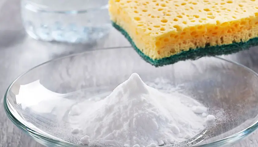 Ein Berg weißes Pulver, das ist Soda, liegt in einer Glasschale. Daneben ein Schwamm und ein Glas Wasser.