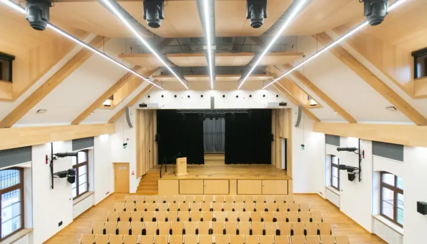 Nowa aula z drewnianymi elementami, sceną i rzędami krzeseł. Idealne miejsce na wykłady, koncerty i spotkania.