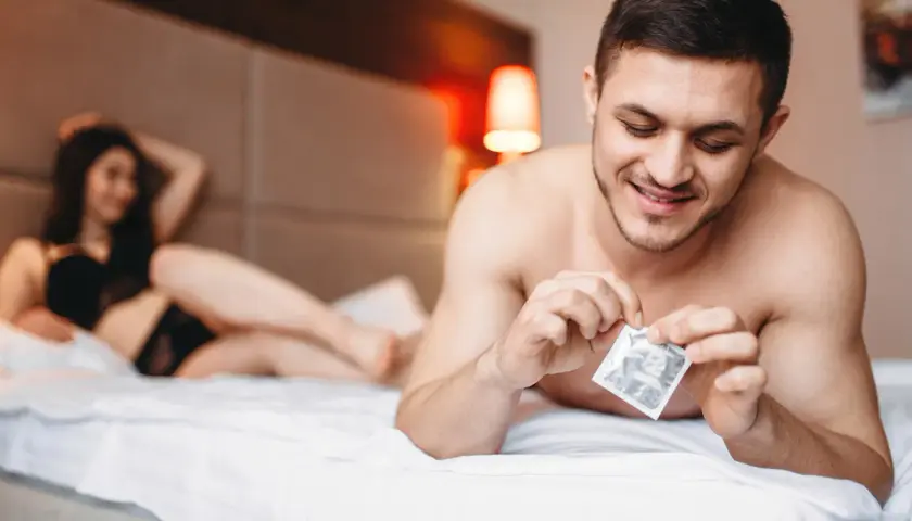 Mężczyzna w łóżku przygotowuje się do intymnego spotkania, trzymając w dłoni prezerwatywę. Kobieta leży w tle. Rozmiary prezerwatyw są kluczowe dla komfortu.