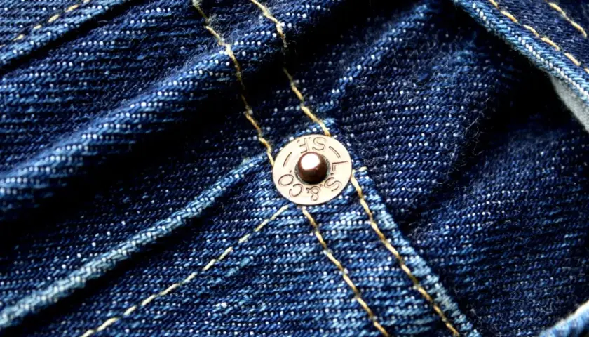 Close-up of dark blue denim with yellow stitching. A copper rivet, marked "S.F. & CO. L.S.", is visible, a testament to the durability of these jeans.