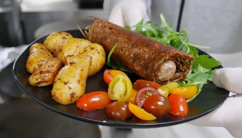 Une galette saucisse roulée, servie avec des pommes de terre rôties et des tomates cerises colorées, sur une assiette noire.