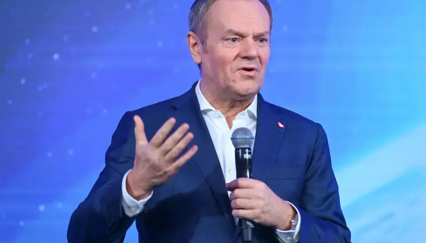 Donald Tusk w garniturze, przemawiający do mikrofonu na tle niebieskiego ekranu.
