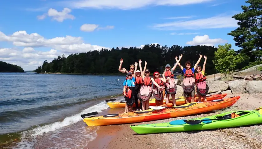 Grupa przyjaciół w kamizelkach ratunkowych pozuje przy kajakach na plaży. W tle widać wodę i zalesione wyspy archipelagu sztokholmskiego.