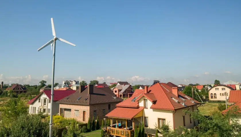 Biała turbina wiatrowa na tle nieba i domów jednorodzinnych.