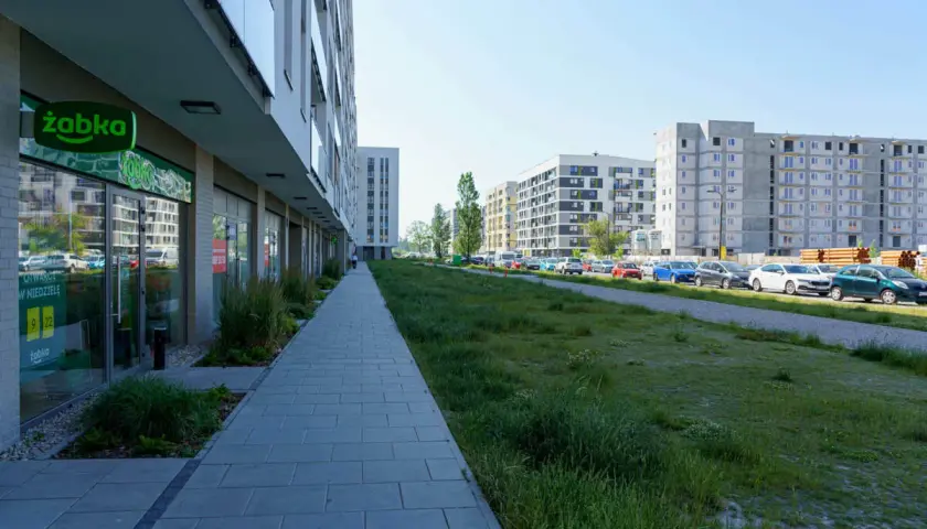 Nowoczesne osiedle w Ursusie Centralnym. Chodnik prowadzi do sklepu Żabka, a dalej widać parking z samochodami i budynki mieszkalne.