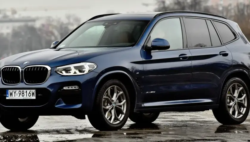 Ciemnoniebieskie BMW X3 na mokrej nawierzchni. Eleganckie linie i sportowy charakter tego SUV-a robią wrażenie.