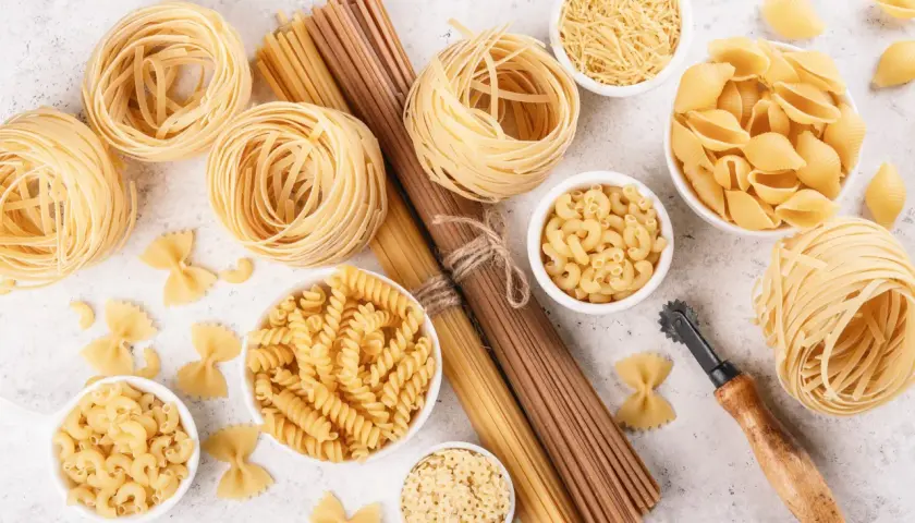 Różnorodne rodzaje makaronu: tagliatelle, fusilli, muszelki, gwiazdki, farfalle i inne, ułożone na jasnym tle.