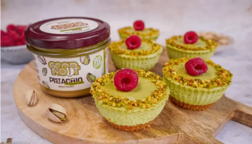 Sześć mini serniczków z kremem pistacjowym, posypanych pistacjami i ozdobionych maliną. Obok słoik kremu pistacjowego.