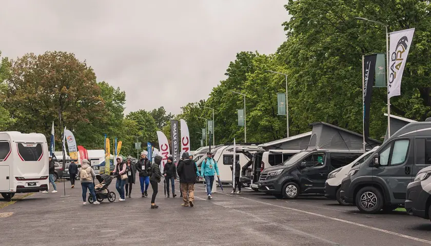 Ludzie zwiedzają wystawę kamperów i przyczep. W tle drzewa i flagi. To zlot caravaningowy.