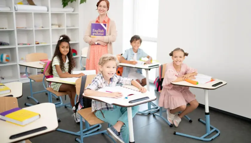 Uśmiechnięta nauczycielka z książką "English grammar" i dzieci przy ławkach. Widać, że to świetny moment, kiedy najlepiej się uczyć.