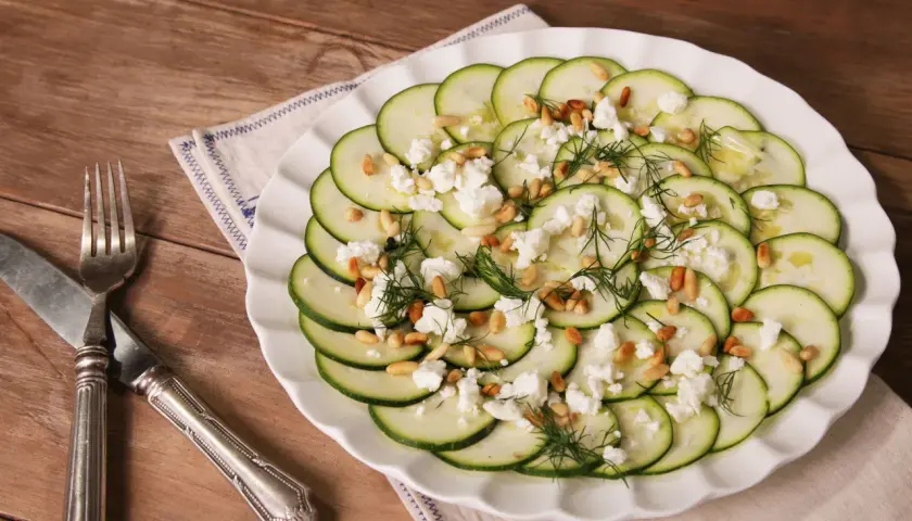 Delicioso carpaccio de calabacín con queso feta desmenuzado, piñones tostados y eneldo fresco, servido en un plato blanco sobre una mesa de madera.