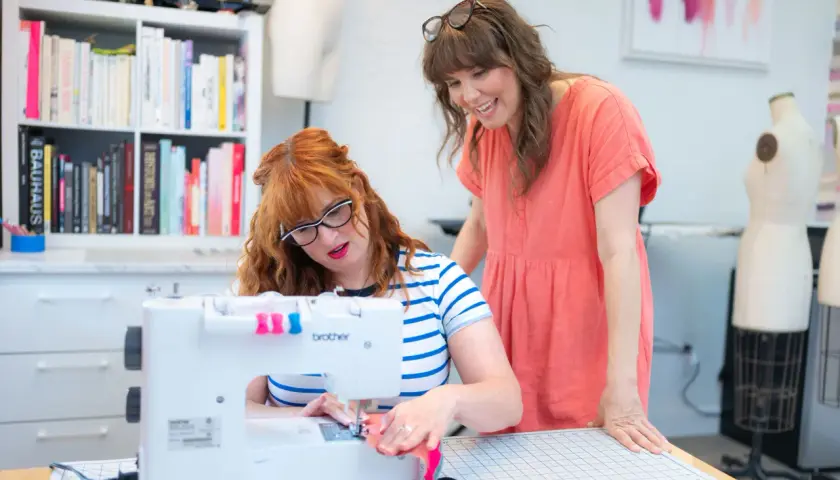 Two women are focused on sewing seams on a white Brother sewing machine in a bright studio.