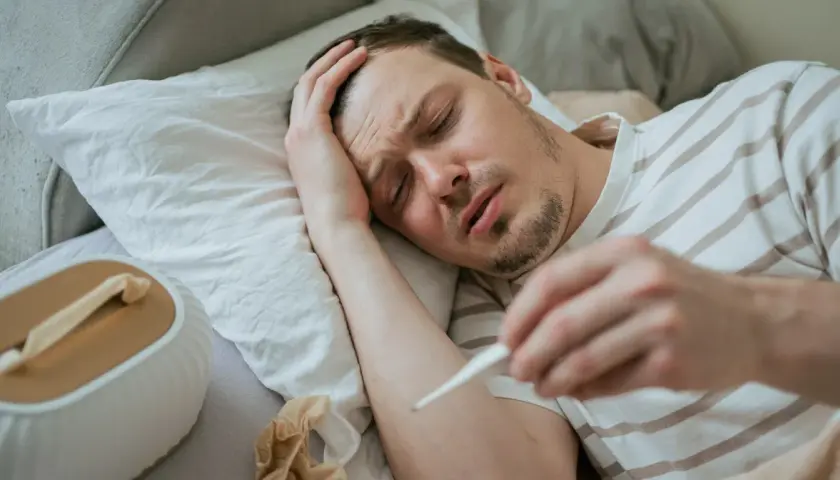 Chory mężczyzna leży w łóżku, trzymając termometr. Widać jego cierpiącą twarz i rękę na czole. To typowe objawy grypy wariant k.