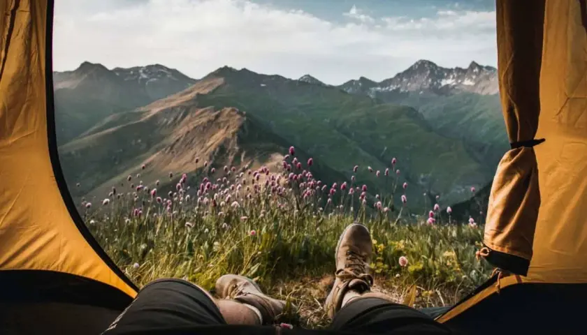 Vista desde una tienda de campaña: piernas con botas de montaña, flores silvestres y montañas verdes bajo un cielo nublado. Disfrutando del acampar con tienda de campaña.