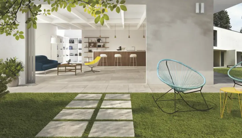 Un sentiero in lastre quadrate su prato verde conduce a un'area living con pavimentazione esterna moderna, che si apre su un interno luminoso con cucina e soggiorno.