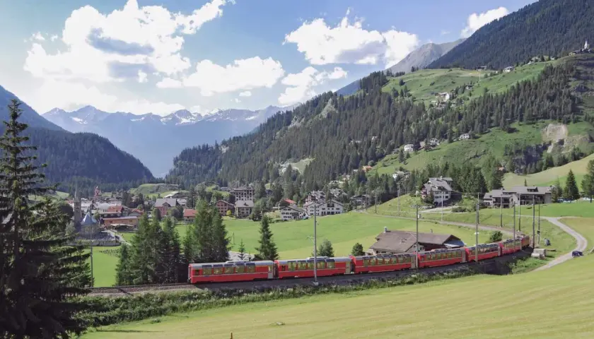Ein roter Zug fährt durch eine malerische Alpenlandschaft. Ein perfekter tagesausflug mit der bahn, vorbei an grünen Wiesen, Wäldern und einem Dorf mit schneebedeckten Bergen im Hintergrund.
